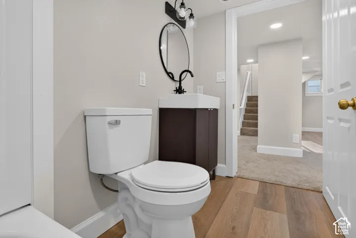 Wood-finish flooring, white toilet, dark wood-tone vanity with a vessel sink, round wall mirror, and matte black lighting fixture