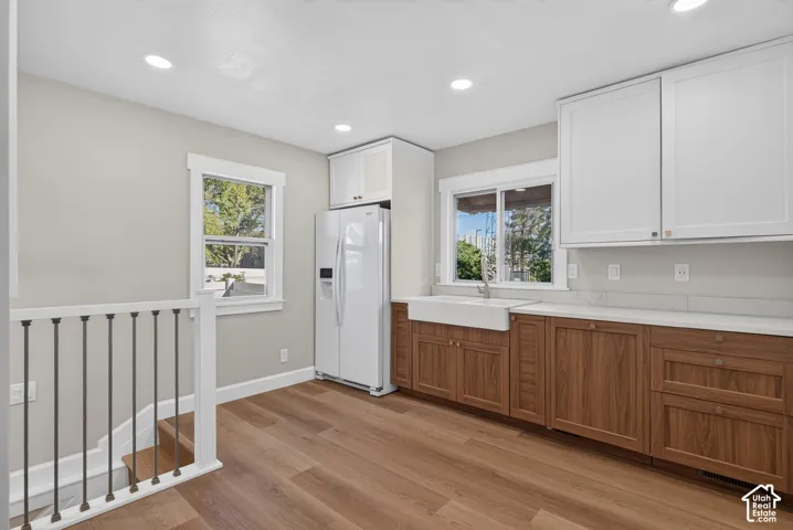 Wood-finish flooring, two-tone cabinetry with white upper cabinets and wood-finish lower cabinets, white farmhouse sink with a chrome faucet, white countertops, and recessed lighting