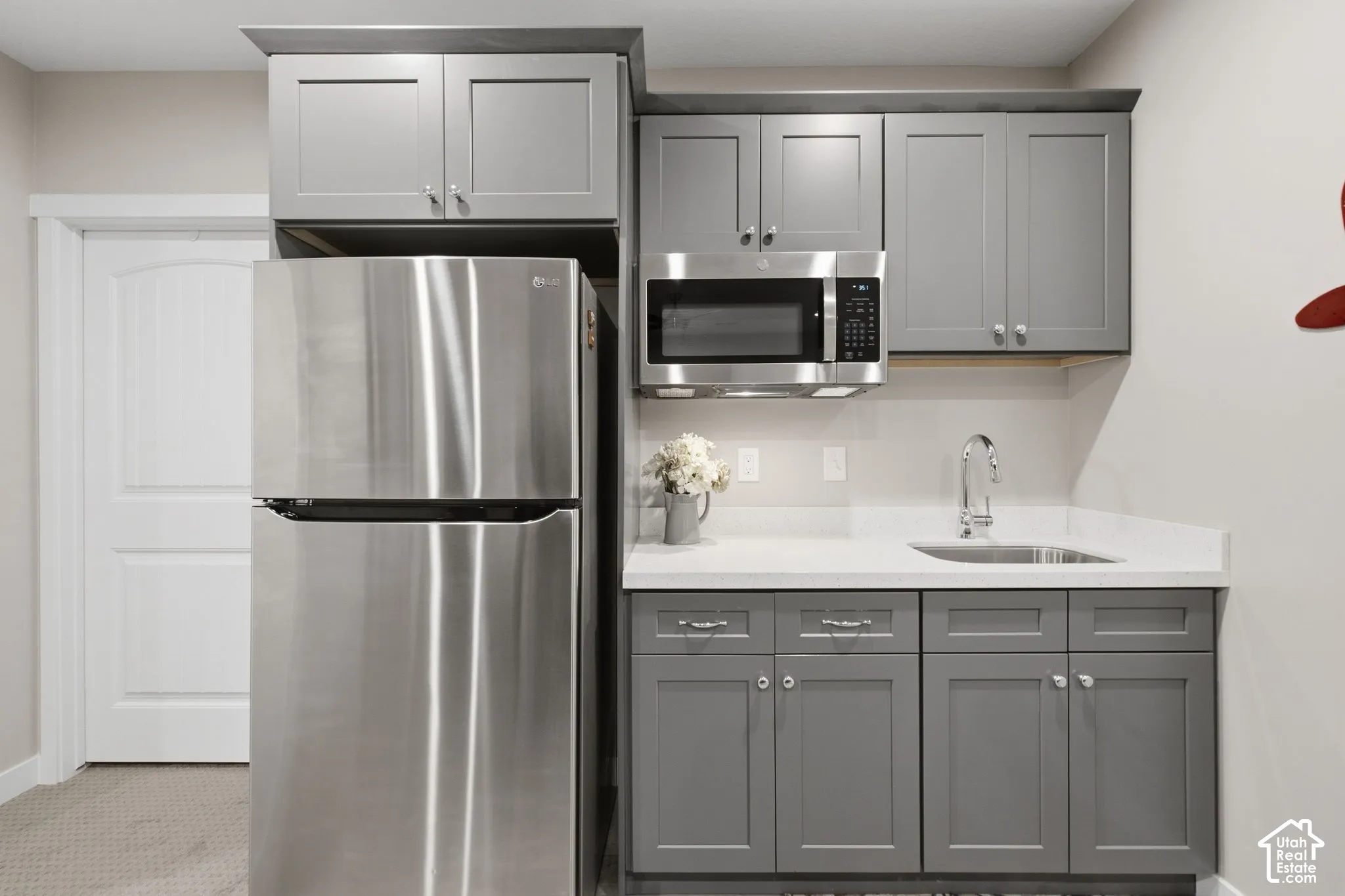 Stainless steel refrigerator, built-in microwave, gray shaker-style cabinetry, white solid surface countertop, and stainless steel sink with a chrome faucet