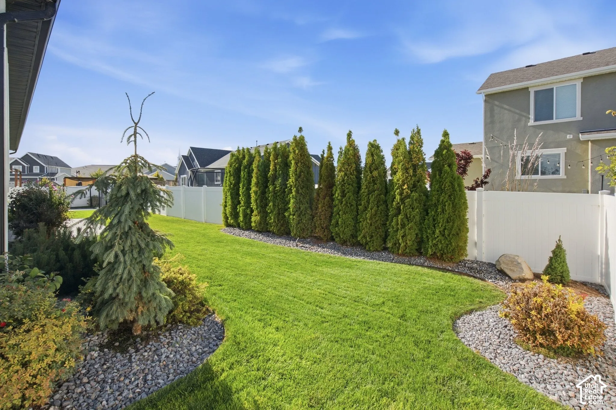 Expansive lawn with a perimeter white vinyl fence, mature evergreen trees, and decorative rock landscaping