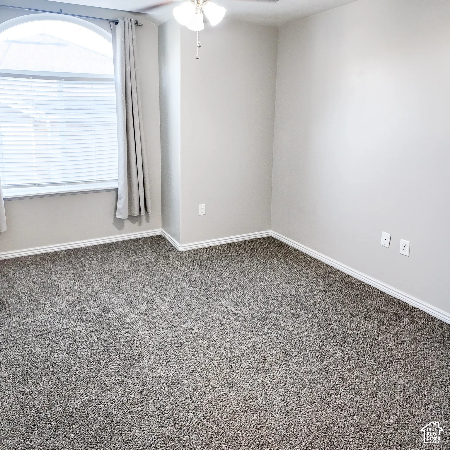 Arched window with blinds, overhead ceiling fan with light fixture, light gray wall paint, and gray carpet flooring
