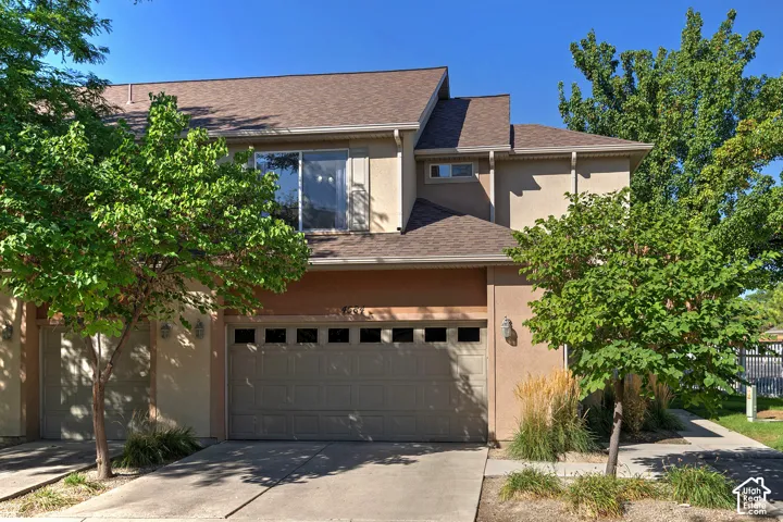 Multi-level stucco exterior with an attached garage, featuring a brown shingle roof and mature landscaping