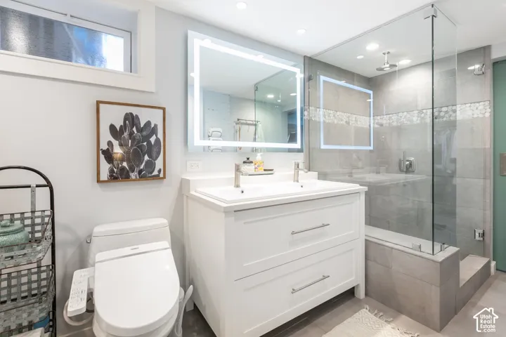 White vanity with dual sinks, an illuminated mirror, a glass-enclosed shower, a rainfall showerhead, and a tiled shower bench