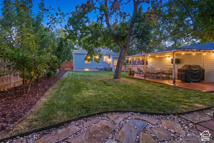 Expansive backyard featuring a stone patio, covered outdoor living area, established trees, and a manicured lawn