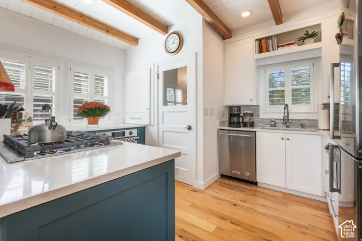 Kitchen featuring wood-finish flooring, exposed wood ceiling beams, white cabinetry, integrated stainless steel appliances, and a central island with a gas range and light-toned countertop