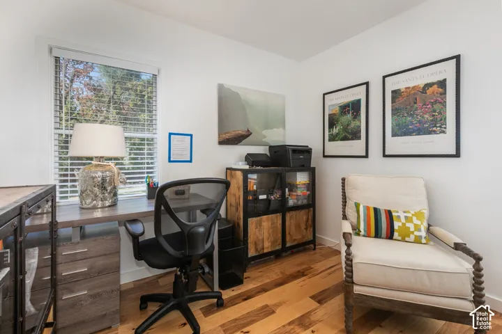 Hardwood flooring, crisp white walls, single window with blinds, built-in desk with drawers, and a storage cabinet with wood-finish doors