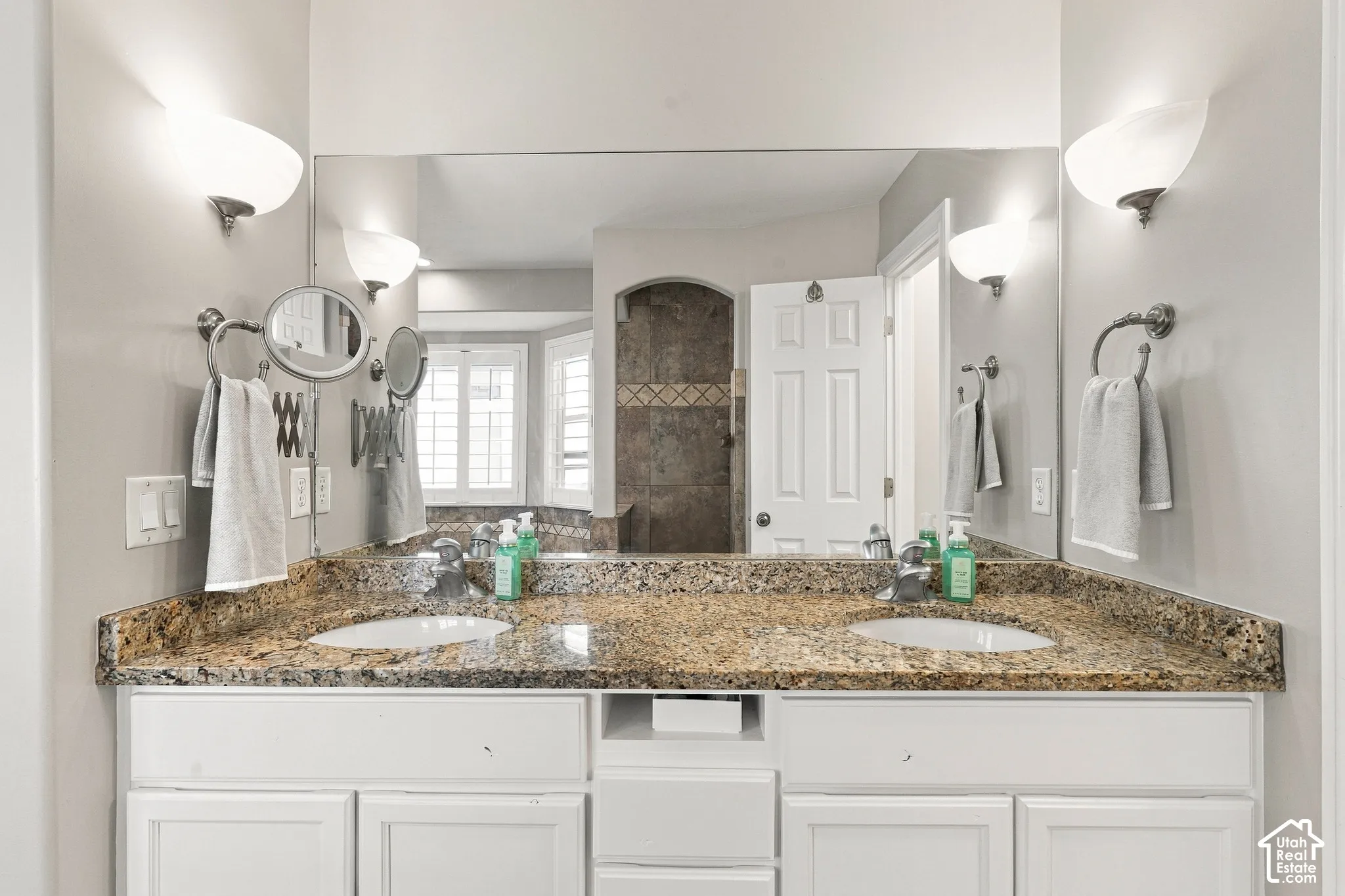 Double vanity with granite countertop, integrated sinks, white cabinetry, and wall-mounted sconces