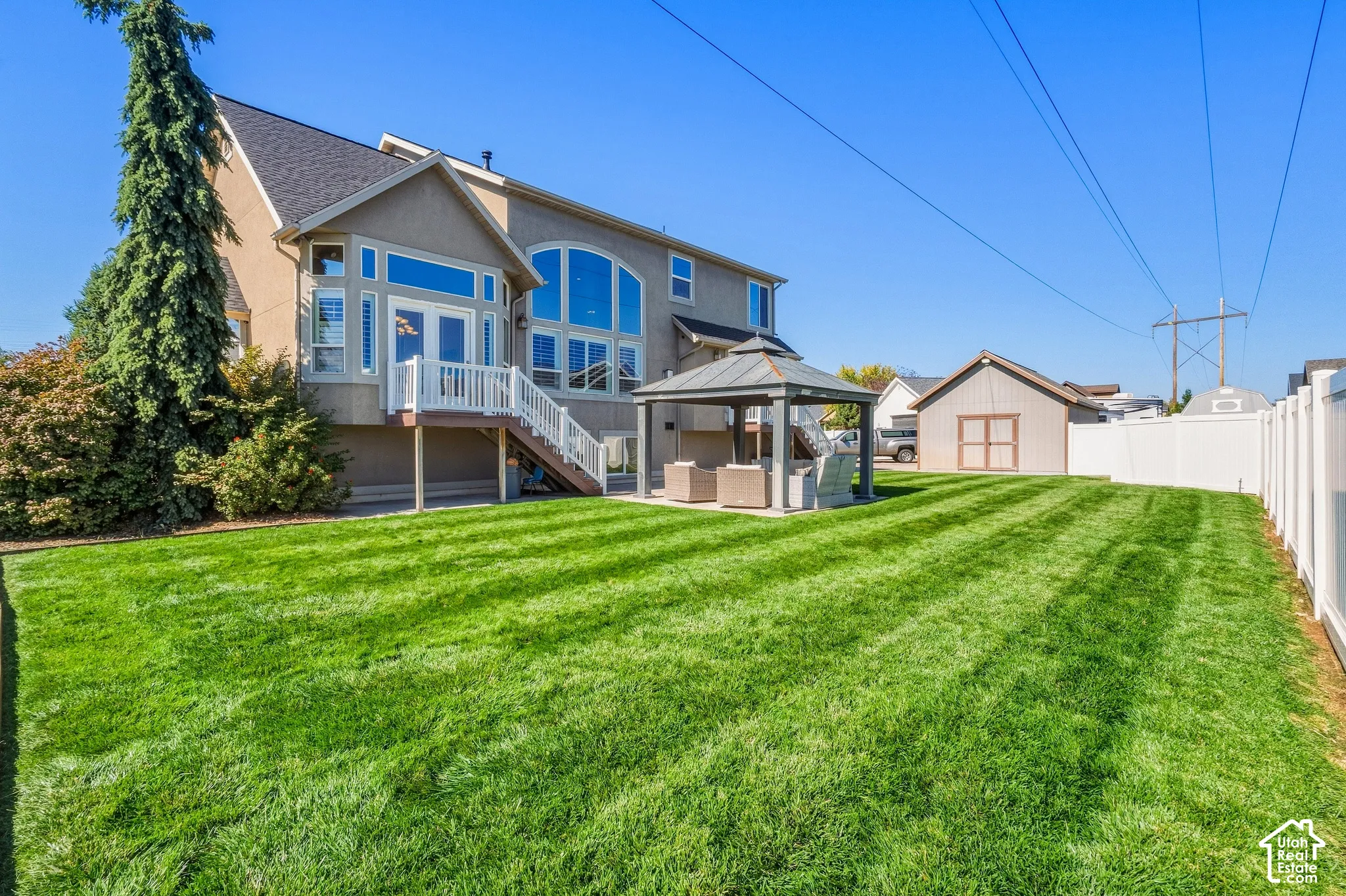 Expansive green lawn, detached storage shed, covered patio structure, white privacy fencing, and two-story exterior with large windows