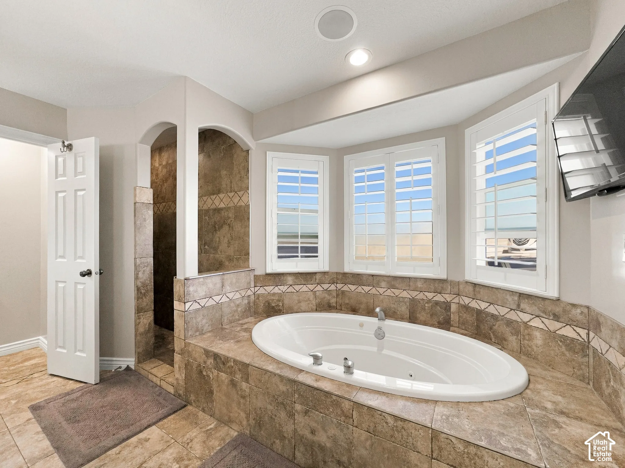 Soaking tub set in a tiled surround with decorative accents, an arched walk-in shower, tiled flooring, and three bay windows with plantation shutters