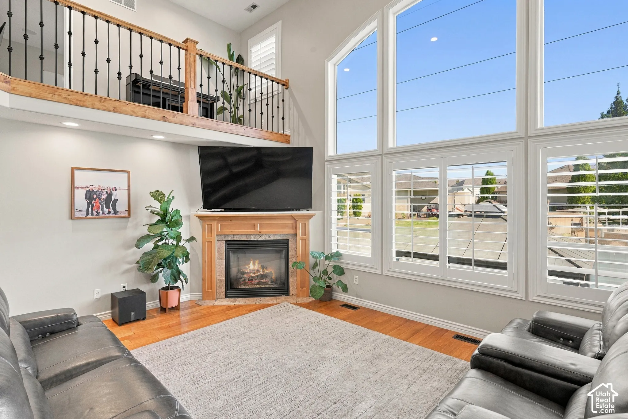 Two-story living room featuring a gas fireplace with a wood mantel, a second-story loft with an iron baluster railing, large arched windows with plantation shutters, and wood-finish flooring