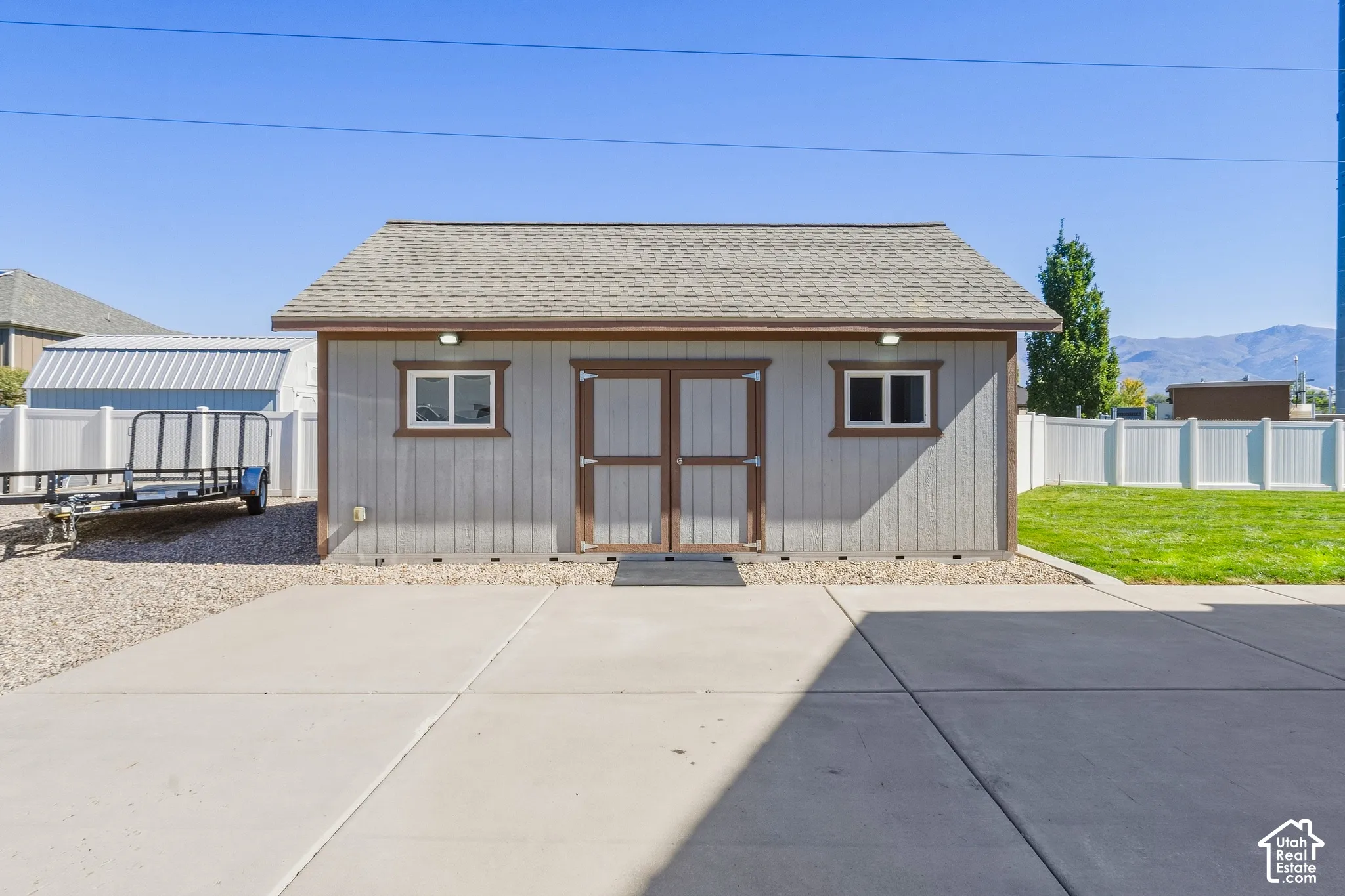 Detached shed with gabled roof, panel siding, double entry doors, two windows, and exterior lighting, bordered by a concrete pad and gravel