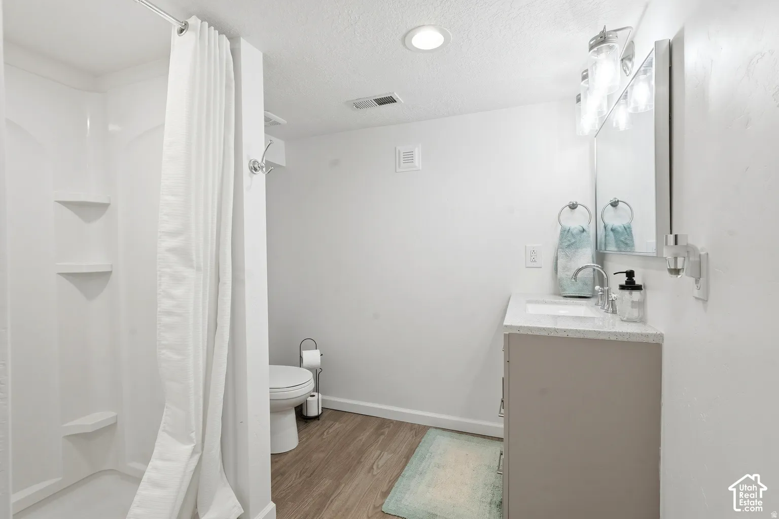 Shower with built-in shelving, wood-finish flooring, single vanity with light-toned countertop, wall-mounted mirror, and overhead lighting