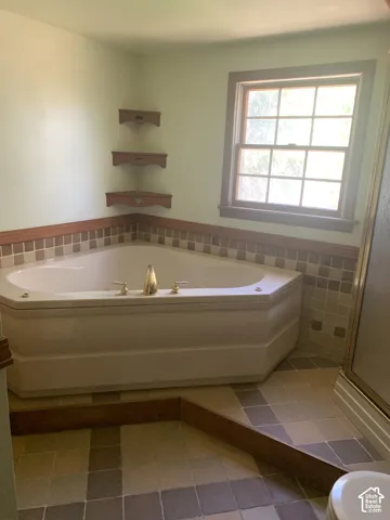 Corner soaking tub with brass fixtures, wood-framed window, and tiled accent wall