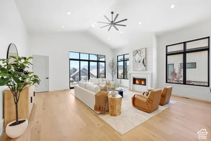 Vaulted ceiling with a contemporary ceiling fan, wide plank wood-finish flooring, a fireplace with a white mantel and shiplap-style accent wall, and large black-framed windows