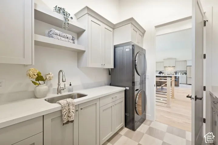 Laundry room featuring stacked washer and dryer, stainless steel sink, light-toned cabinetry, open shelving, and light-toned flooring