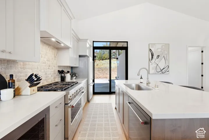 Vaulted ceiling kitchen featuring white cabinetry, a stone tile backsplash, a stainless-steel range with hood, and a large island with a sink