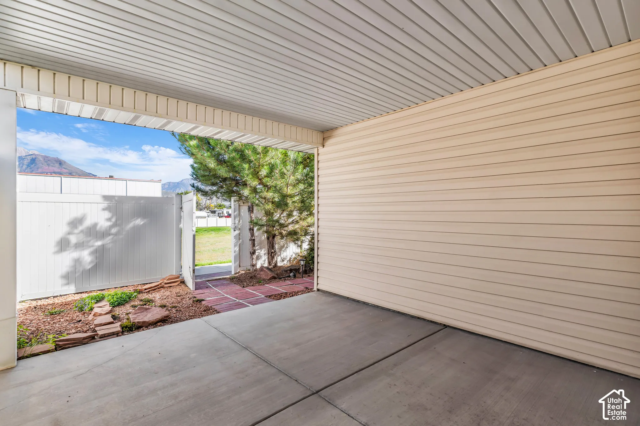 Covered patio with concrete flooring, beige siding, white vinyl fencing, and brick paver pathway
