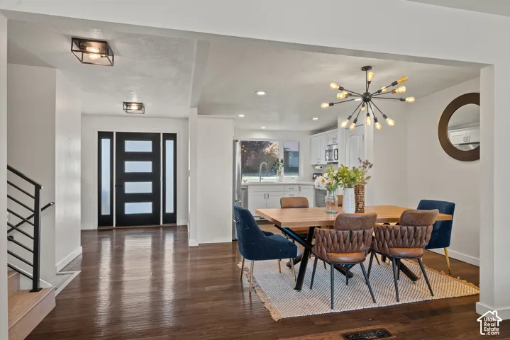 Open-concept dining and kitchen area showcasing dark wood-finish flooring, a linear chandelier with brass accents, recessed ceiling lighting, and a modern black-frame entry door with frosted glass panels