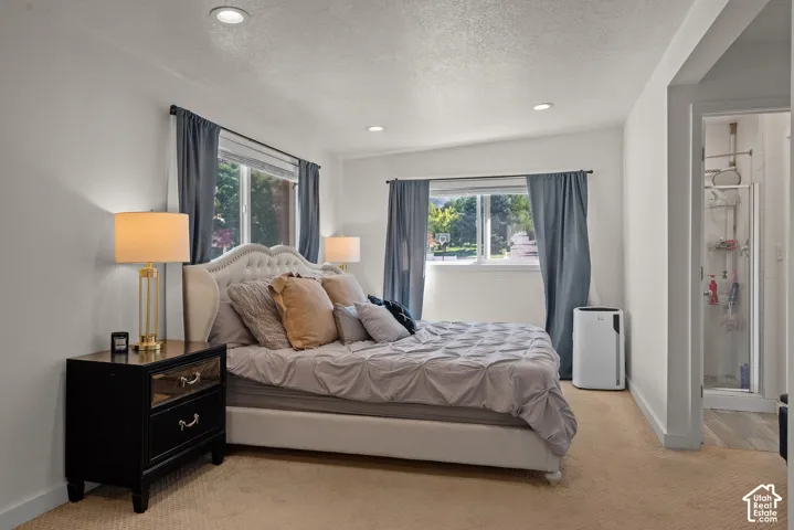 Bright primary bedroom showcasing dual windows with blue drapery, recessed ceiling lighting, warm bedside lamps, black nightstand with drawer storage, and an ensuite bathroom accessible through a glass-paneled door