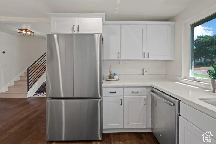 Modern kitchen featuring stainless steel French-door refrigerator with bottom freezer drawer, white shaker-style cabinetry with brushed nickel hardware, quartz countertops, stainless steel dishwasher, and a large window overlooking outdoor landscaping
