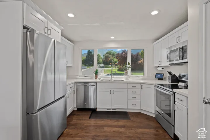 White shaker-style cabinetry with chrome hardware, stainless steel appliances, wood-finish flooring, recessed lighting, and a corner sink with a large window