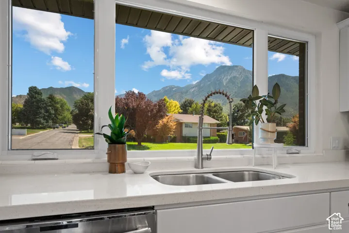 Expansive window framing a mountain backdrop, dual basin stainless steel sink, polished chrome gooseneck faucet, light-toned countertop, and white cabinetry