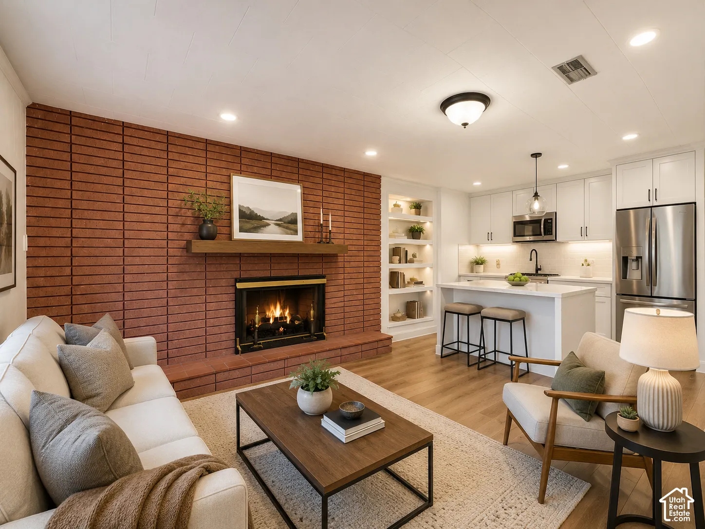 Brick fireplace with wood mantel, built-in shelving with accent lighting, light wood-finish flooring, white cabinetry, and stainless steel appliances