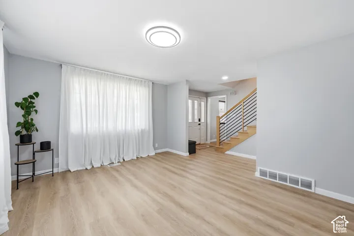 Light wood-finish flooring, white walls, large window with sheer white curtains, modern ceiling light fixture, and a staircase with light wood treads and black horizontal railings