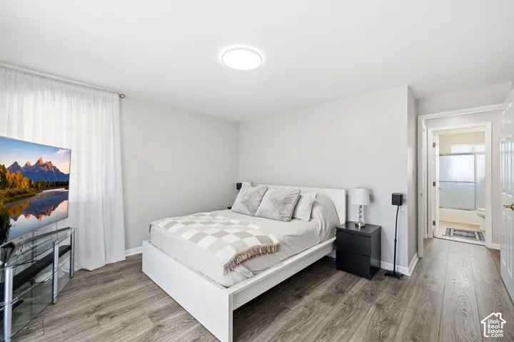 Wood-finish flooring, light neutral wall color, white window coverings, flush-mount ceiling light, and an adjoining bathroom with a tub/shower combination