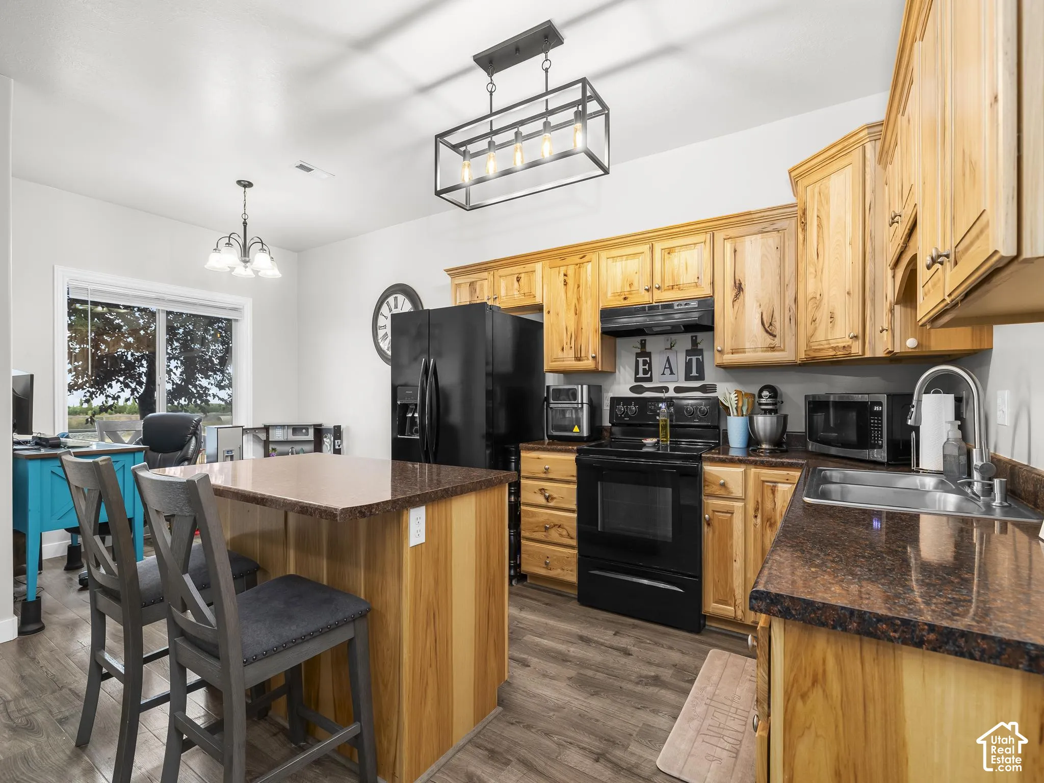 Wood-finish flooring, wood cabinetry, dark countertops, built-in stainless steel sink, and black appliances