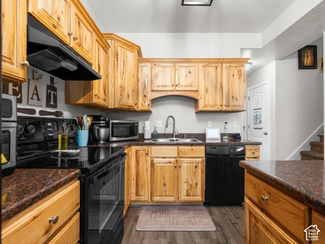 Kitchen featuring wood cabinetry, dark countertops, a stainless steel sink, black appliances, and wood-finish flooring