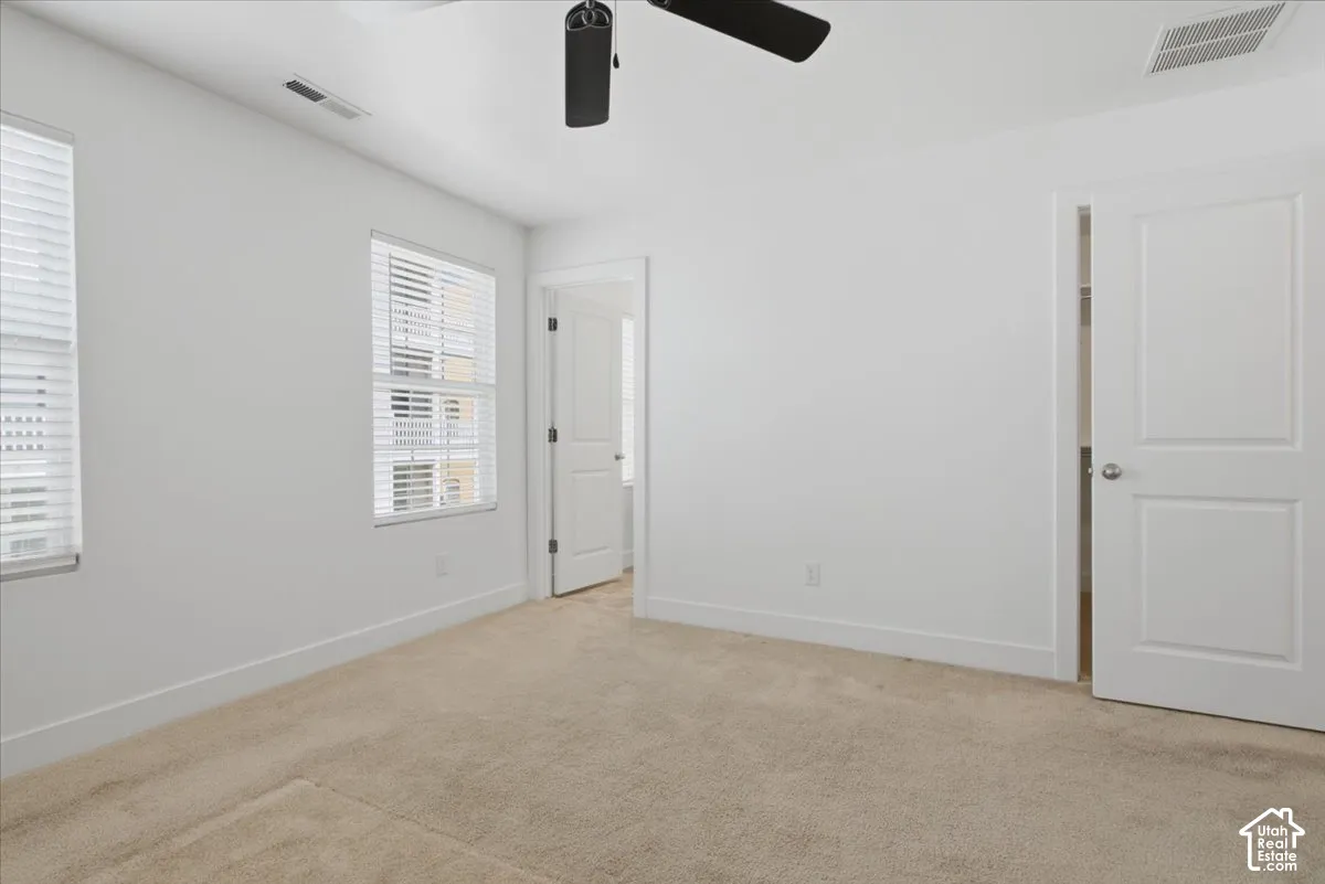 Plush carpet flooring, white painted walls, two windows with blinds, a ceiling fan, and white interior doors