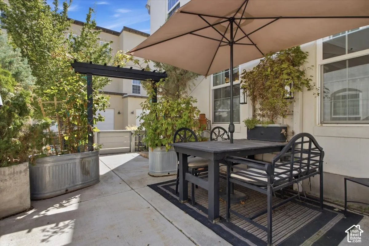 Concrete patio featuring a black pergola, various planted greenery, and exterior wall sconces