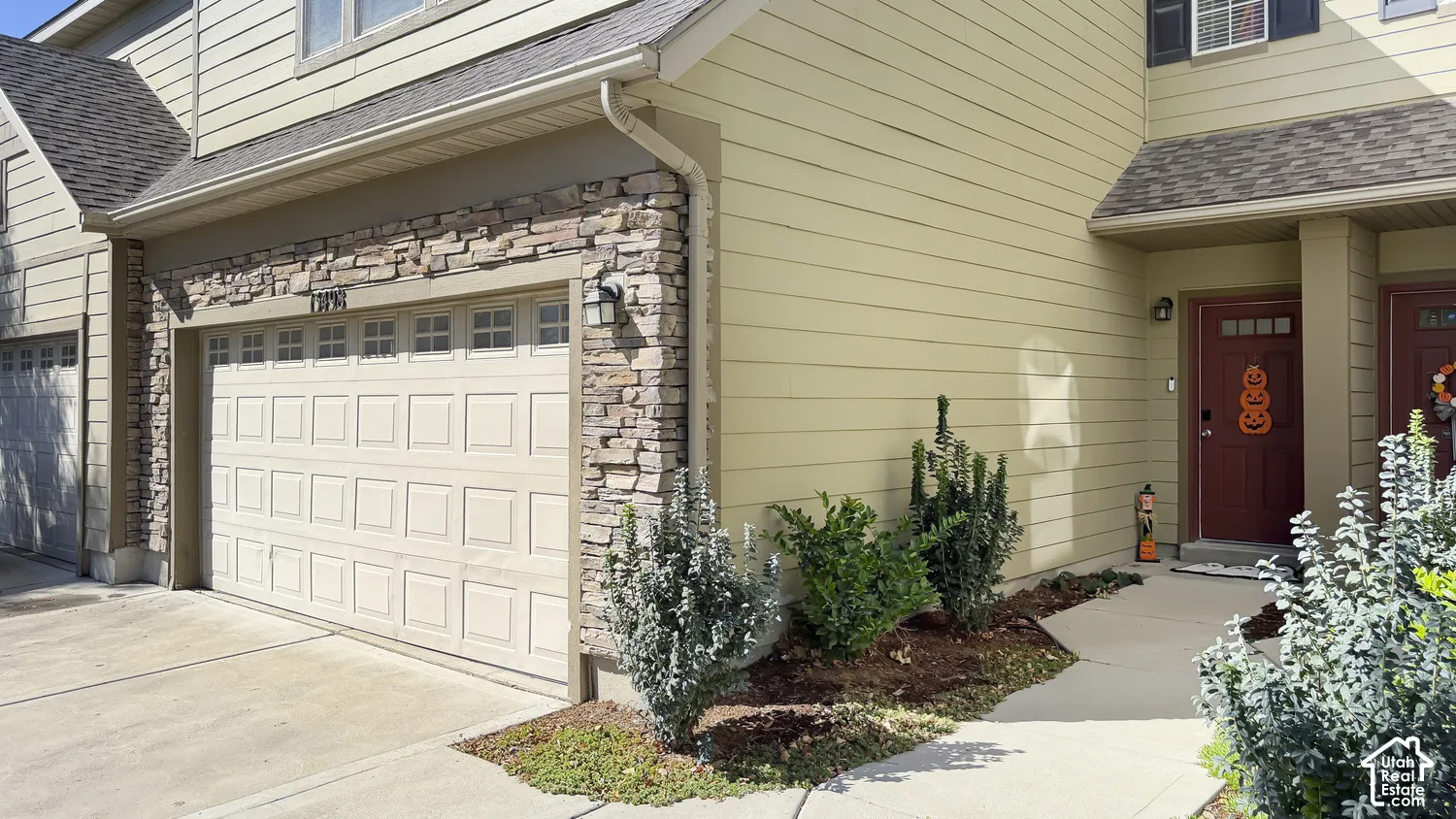 Stone facade accents, attached garage with panel door, concrete driveway, front entry with covered porch, and mature landscaping