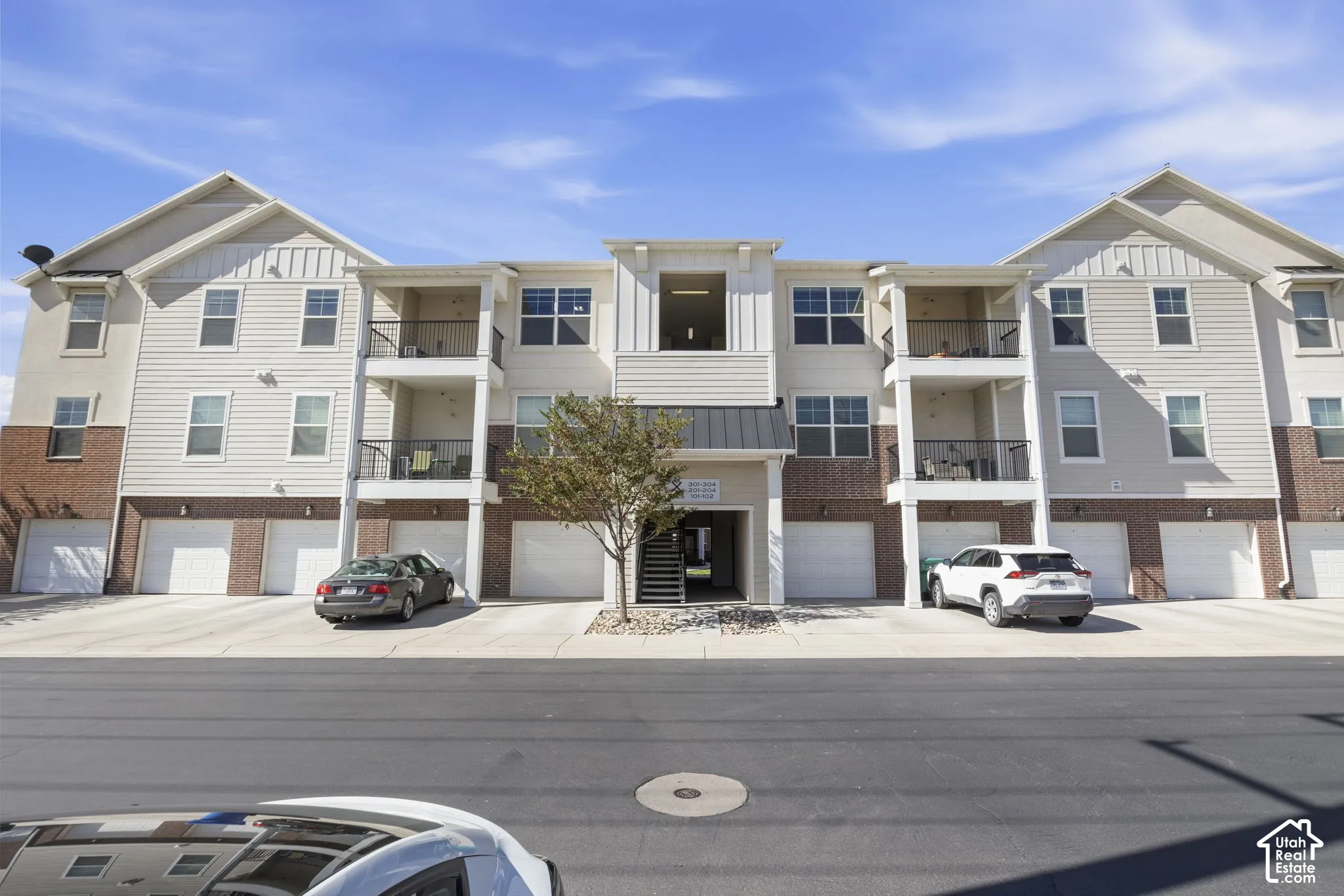 Multi-story building featuring light-toned siding, brick accents, multiple balconies with dark railings, and individual garage doors