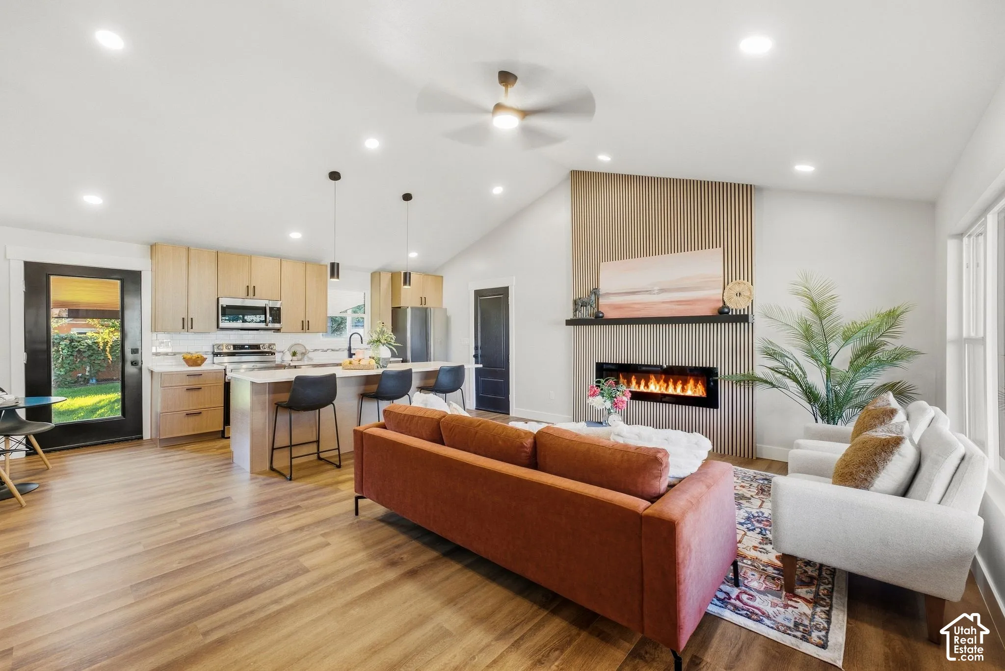 Vaulted ceilings, wood-finish flooring, a contemporary fireplace with a decorative wood slat wall, recessed lighting, and a kitchen island with pendant lighting