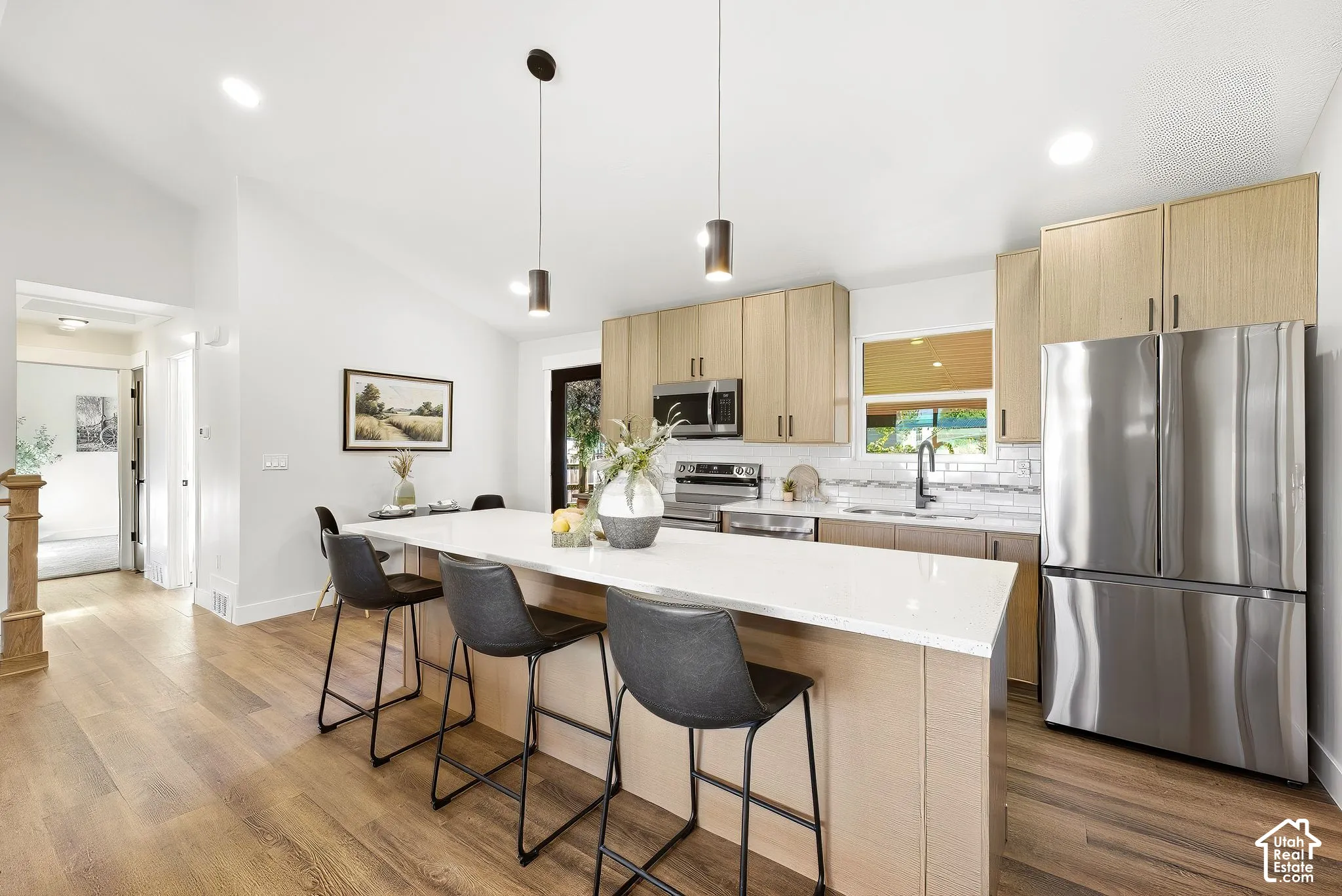Modern kitchen featuring wood-finish flooring, light wood-tone cabinetry, a white countertop island, stainless steel appliances, and a subway tile backsplash