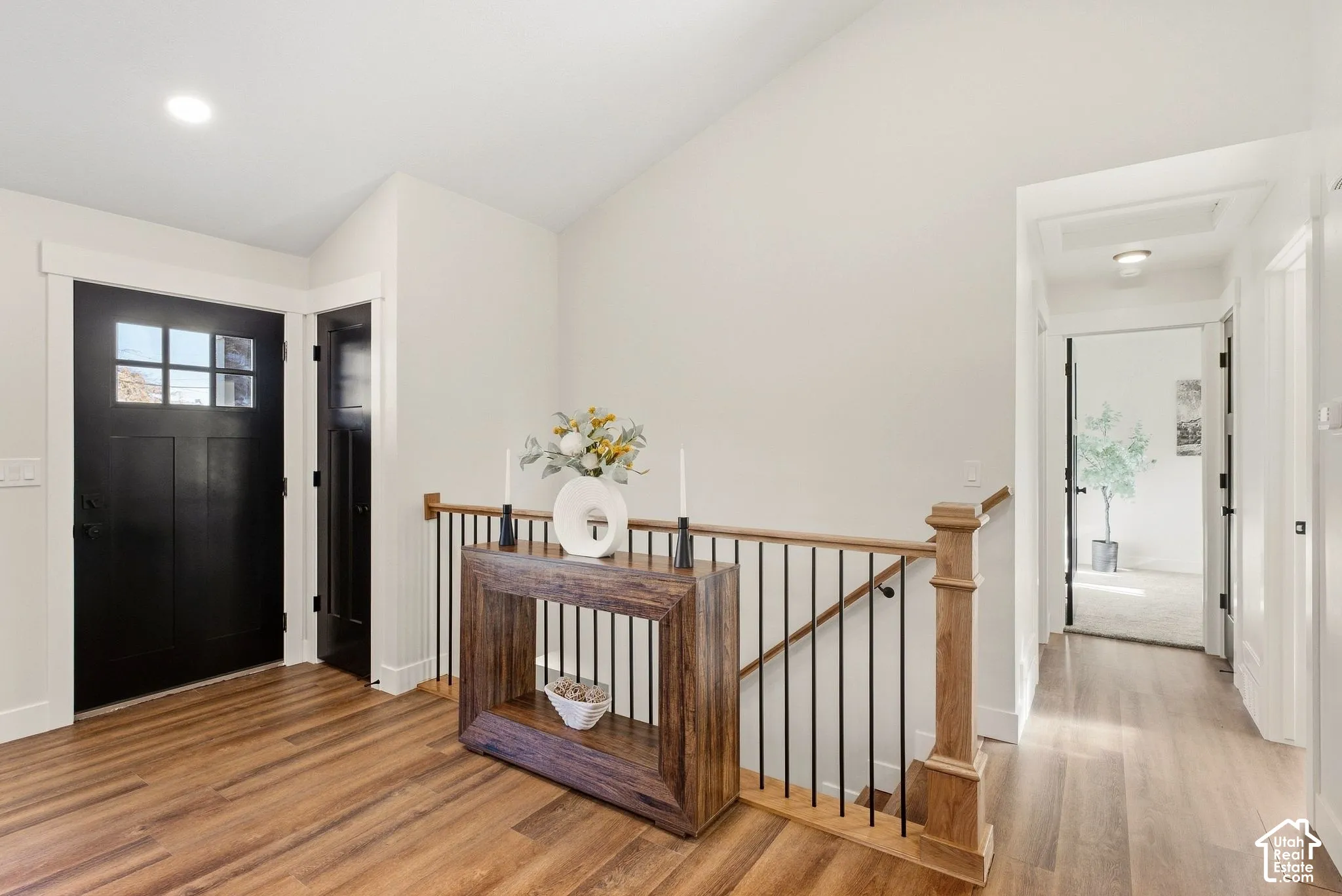 Wood-finish flooring throughout, featuring a craftsman-style entry door with glass inserts, a wood handrail with black metal balusters, and recessed ceiling lighting