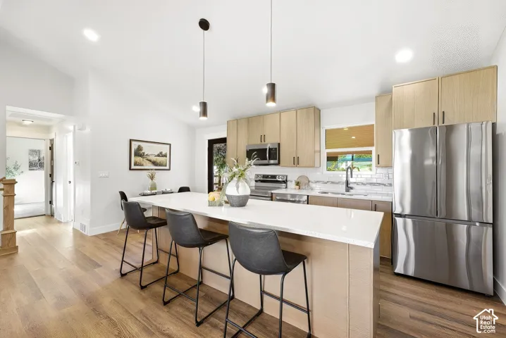 Modern kitchen featuring wood-finish flooring, light wood-tone cabinetry, a white countertop island, stainless steel appliances, and a subway tile backsplash