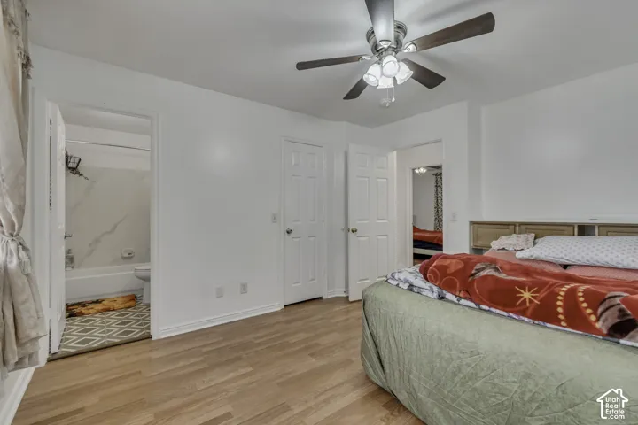 Wood-finish flooring throughout, white walls, ceiling fan with integrated lighting, two panel doors, and a bathroom with a white bathtub and tiled surround