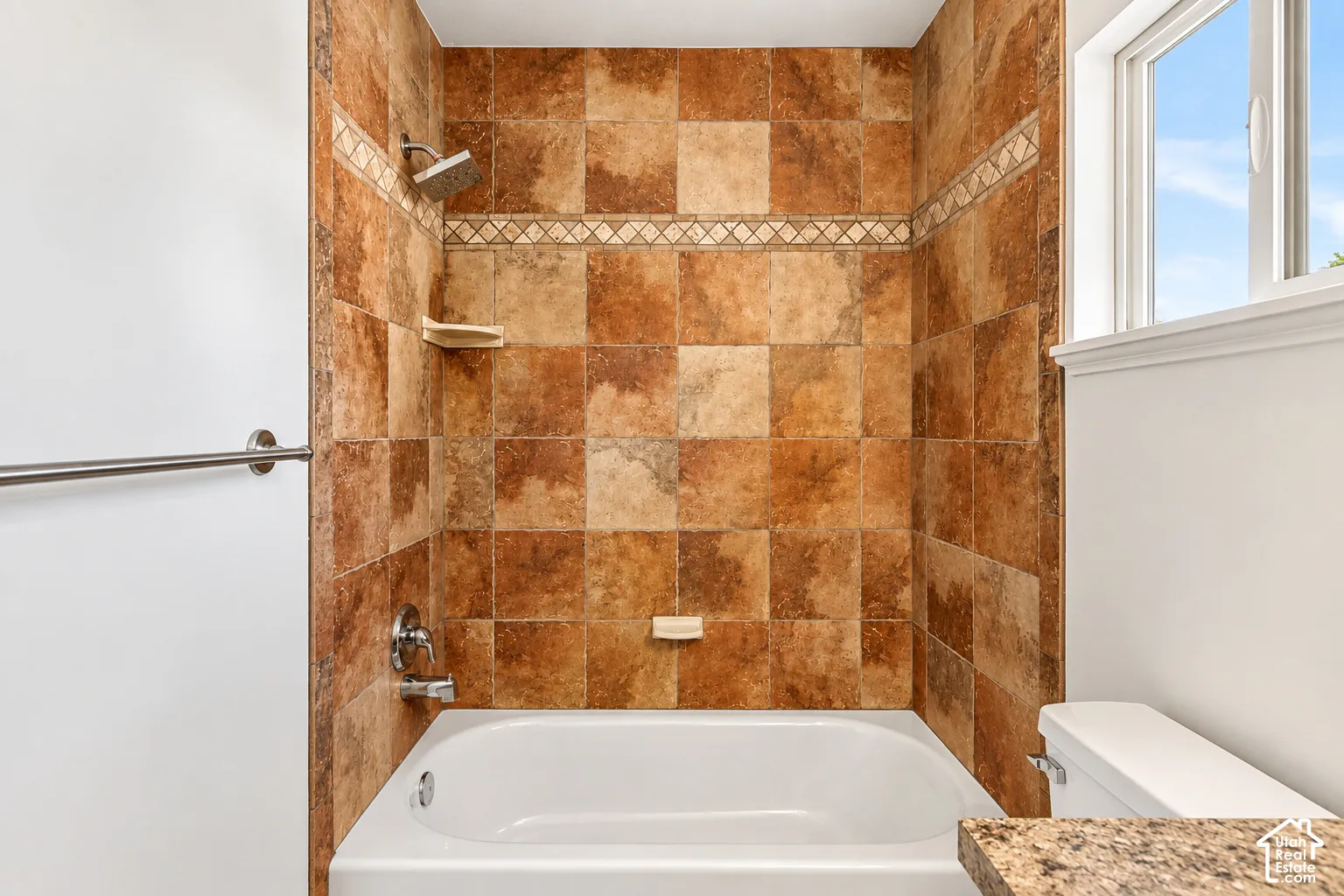Bathtub and shower combination featuring earth-toned wall tile with an accent border, polished chrome fixtures, and a window