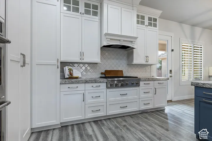 White shaker-style cabinetry, stainless steel range, gray stone countertops, white tile backsplash, and wood-finish flooring