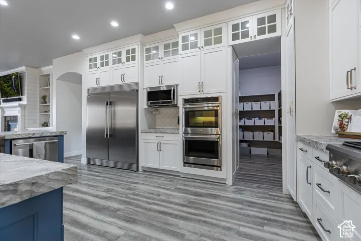 White cabinetry with glass-front uppers, stainless steel double ovens, stainless steel microwave, and wood-finish flooring