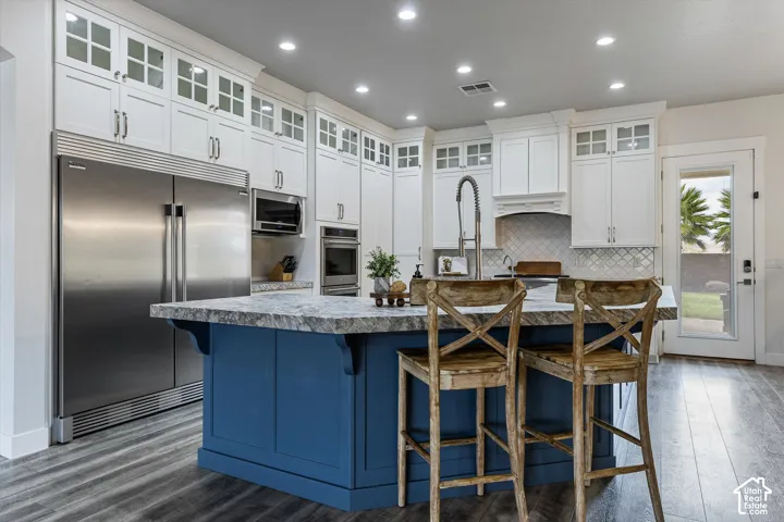 Expansive kitchen featuring a blue island, wood-finish flooring, white cabinetry with glass inserts, stainless steel appliances, and recessed lighting