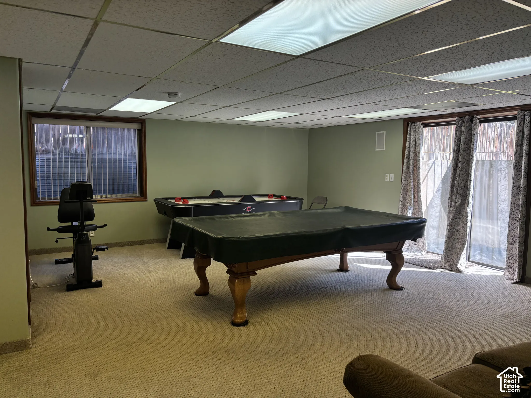 Carpeted entertainment area featuring a drop ceiling with integrated lighting, wood-trimmed egress window, and glass sliding doors