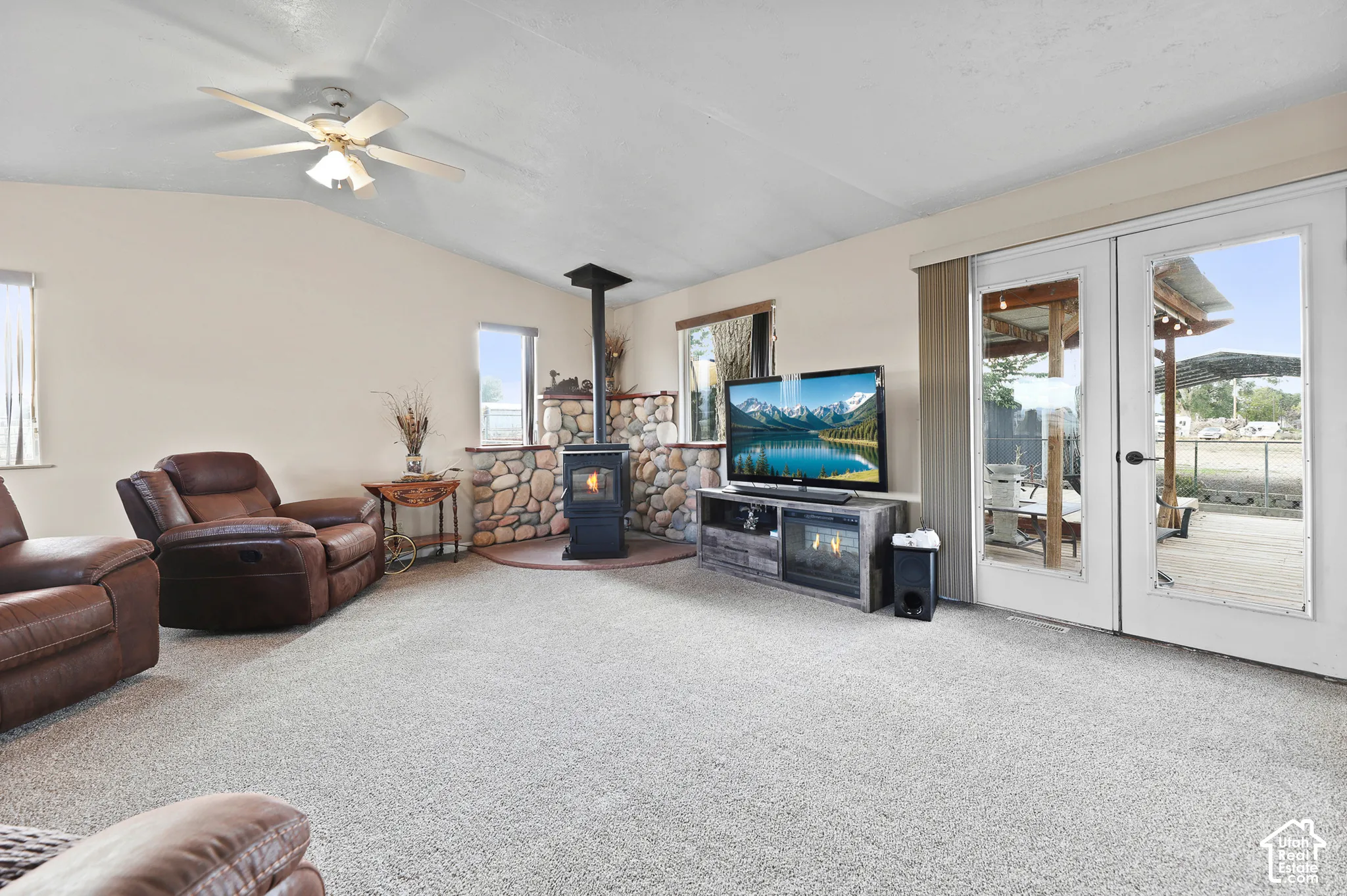 Sloped ceiling with ceiling fan, carpet flooring, stone accent wall, wood-burning stove, and glass-panel double doors opening to a wood deck
