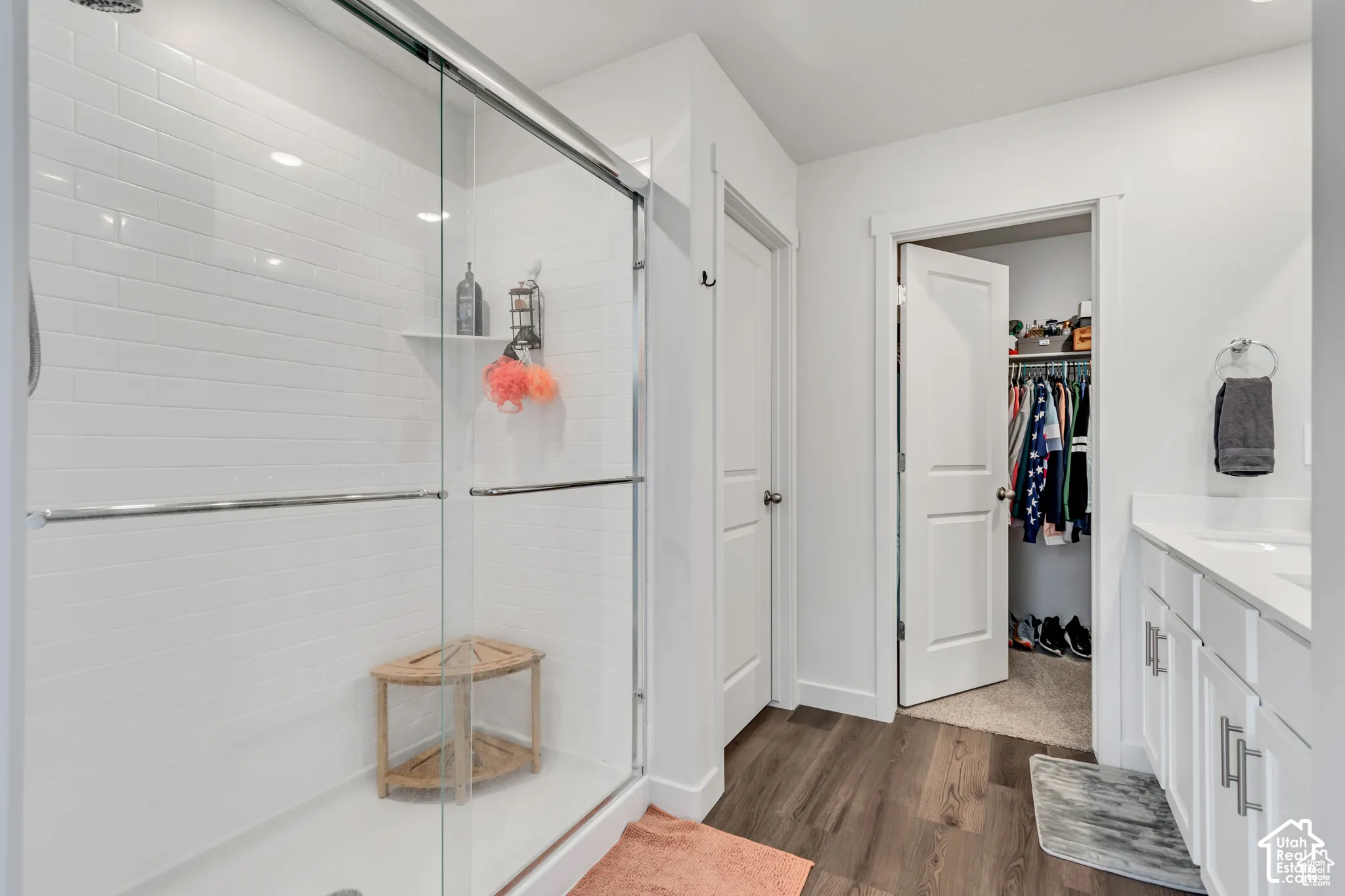 Sliding glass shower enclosure with white subway tile, wood-finish flooring, double vanity with white cabinetry, and white countertops