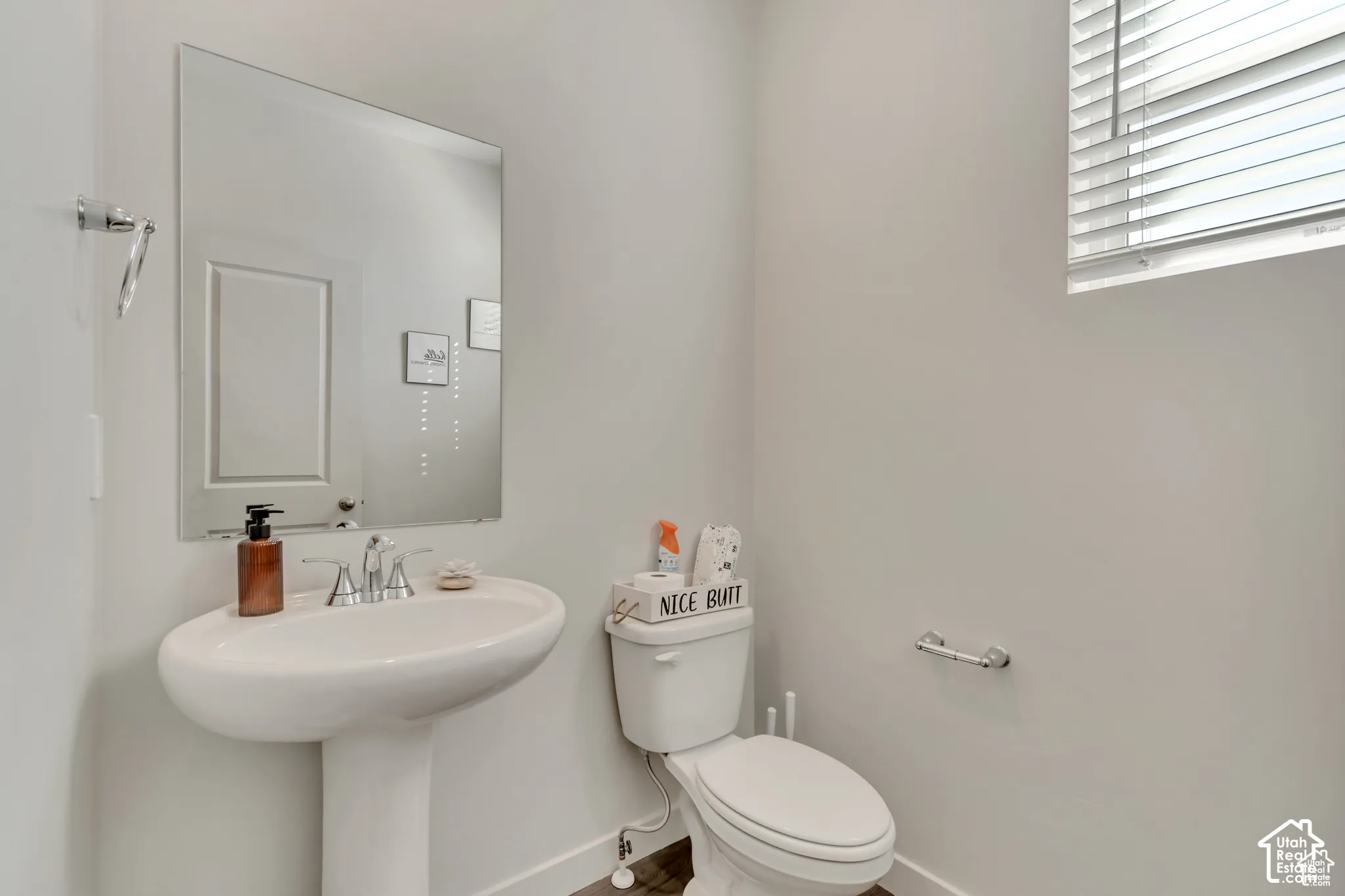 Pedestal sink with chrome faucet, wall-mounted mirror, white toilet, and window with blinds