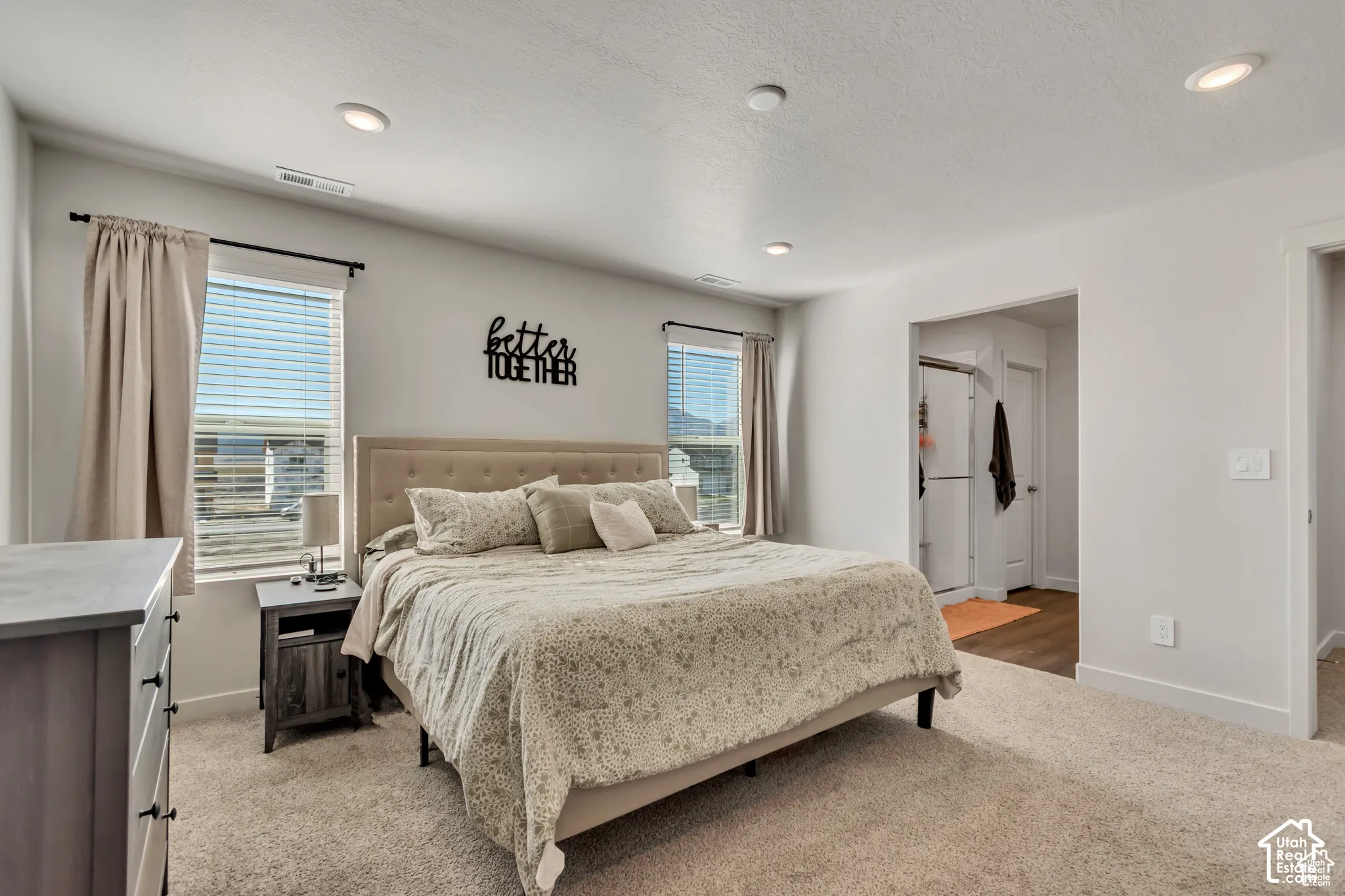 Recessed lighting, light-colored carpeting, two windows with blinds, and an open doorway leading to an adjacent area with wood-finish flooring