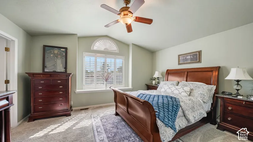 Carpeted flooring, vaulted ceiling, ceiling fan with light fixture, window with plantation shutters, and a decorative arched transom window